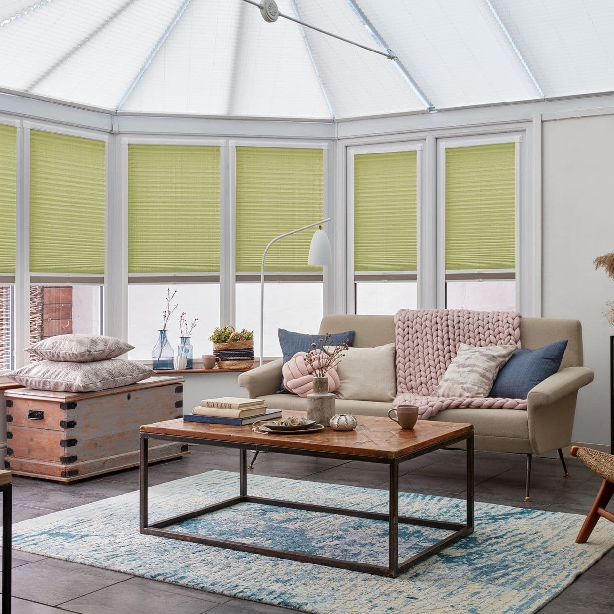 Pleated lime-green cellular blinds, lowered across tall bay windows, filtering soft daylight; in a sunlit conservatory living room with beige sofa, knitted throw, wooden coffee table and blue patterned rug.