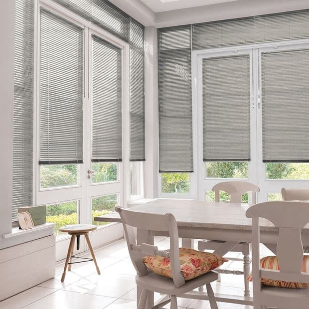 Grey horizontal Venetian blinds lowered across tall French windows, slats closed yet slightly filtering daylight; in a bright sunroom dining area with white table and chairs, garden visible through gaps.