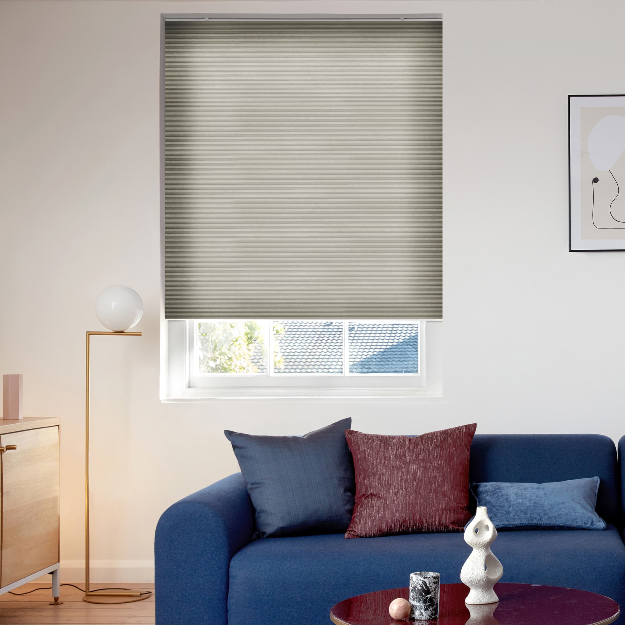 Beige cellular blind, lowered over the window with horizontal ribs and a narrow bottom gap; filtering daylight; above a blue sofa in a living room with a lamp and art.