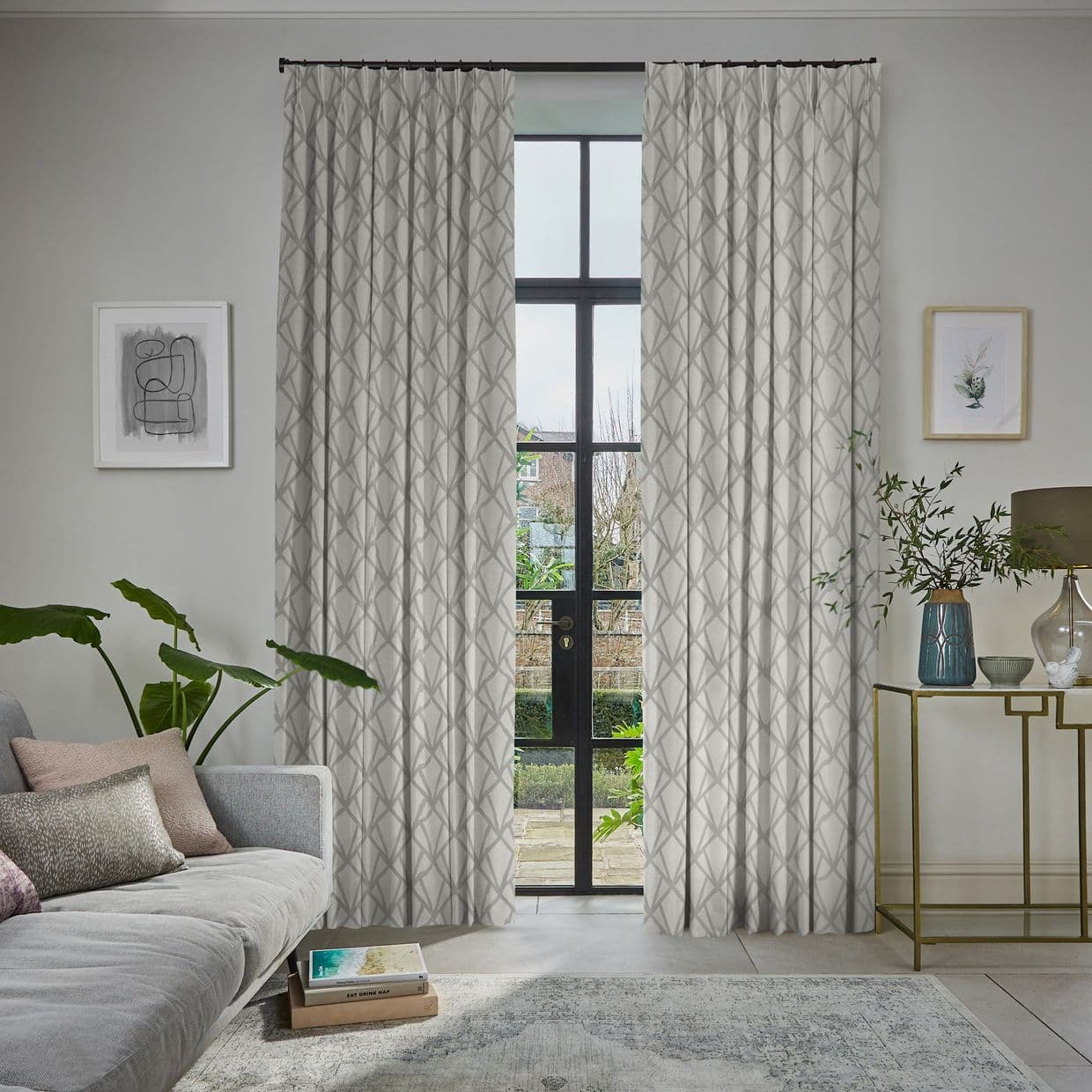 Patterned drapery panels, light grey with a geometric motif, hung on a rod and parted; filtering soft daylight through tall black-framed French doors into a living room overlooking a garden.