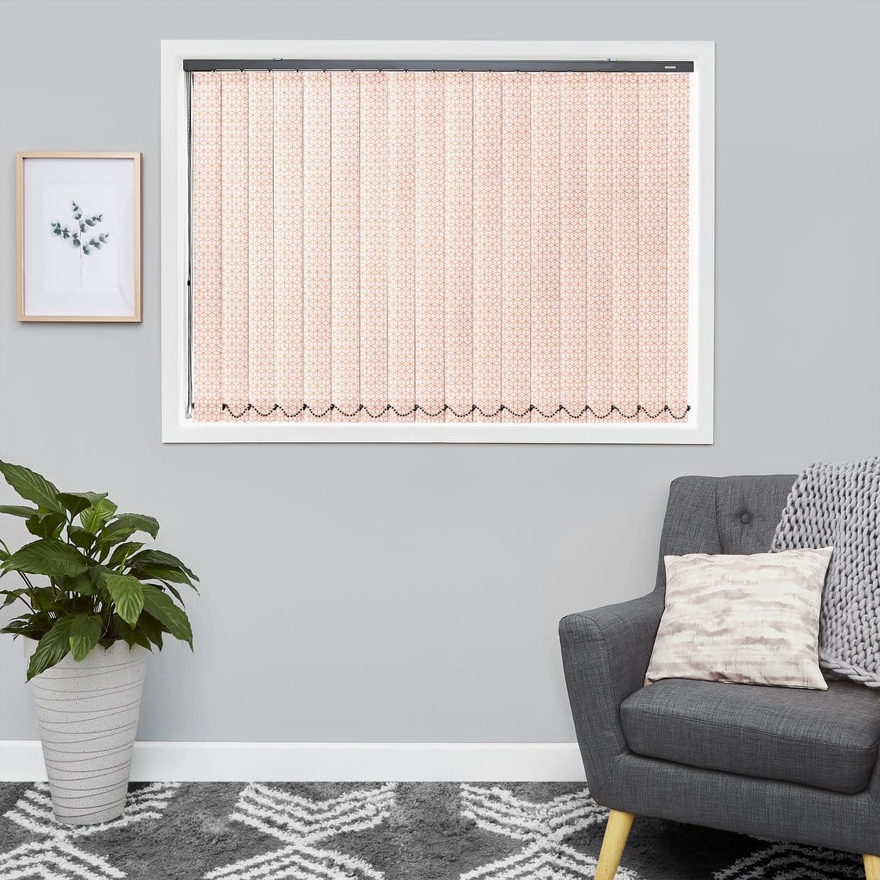 Vertical fabric blinds covering a rectangular window, pale pink geometric pattern, fully closed and filtering soft light; set in a gray living room with armchair, plant, framed art, and rug.