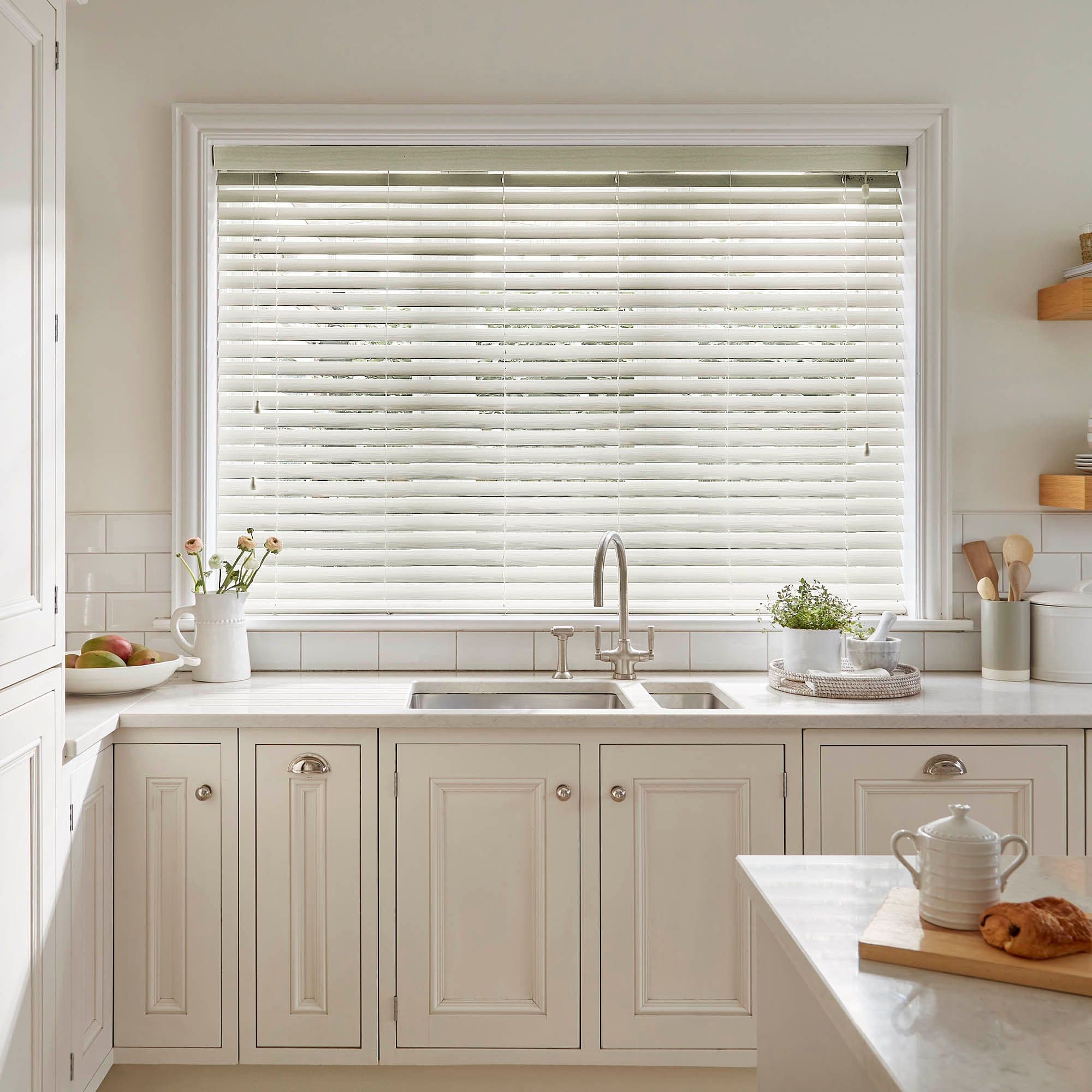 White horizontal Venetian blinds cover a large window, slats mostly closed and softly filtering daylight; above a farmhouse sink in a light, cream kitchen with marble counters, plants, and utensils.