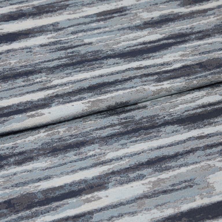 Folded jacquard curtain fabric with horizontal blue-gray, navy, and white striations; draped over itself, revealing woven texture and subtle sheen in a close-up, softly lit studio view.