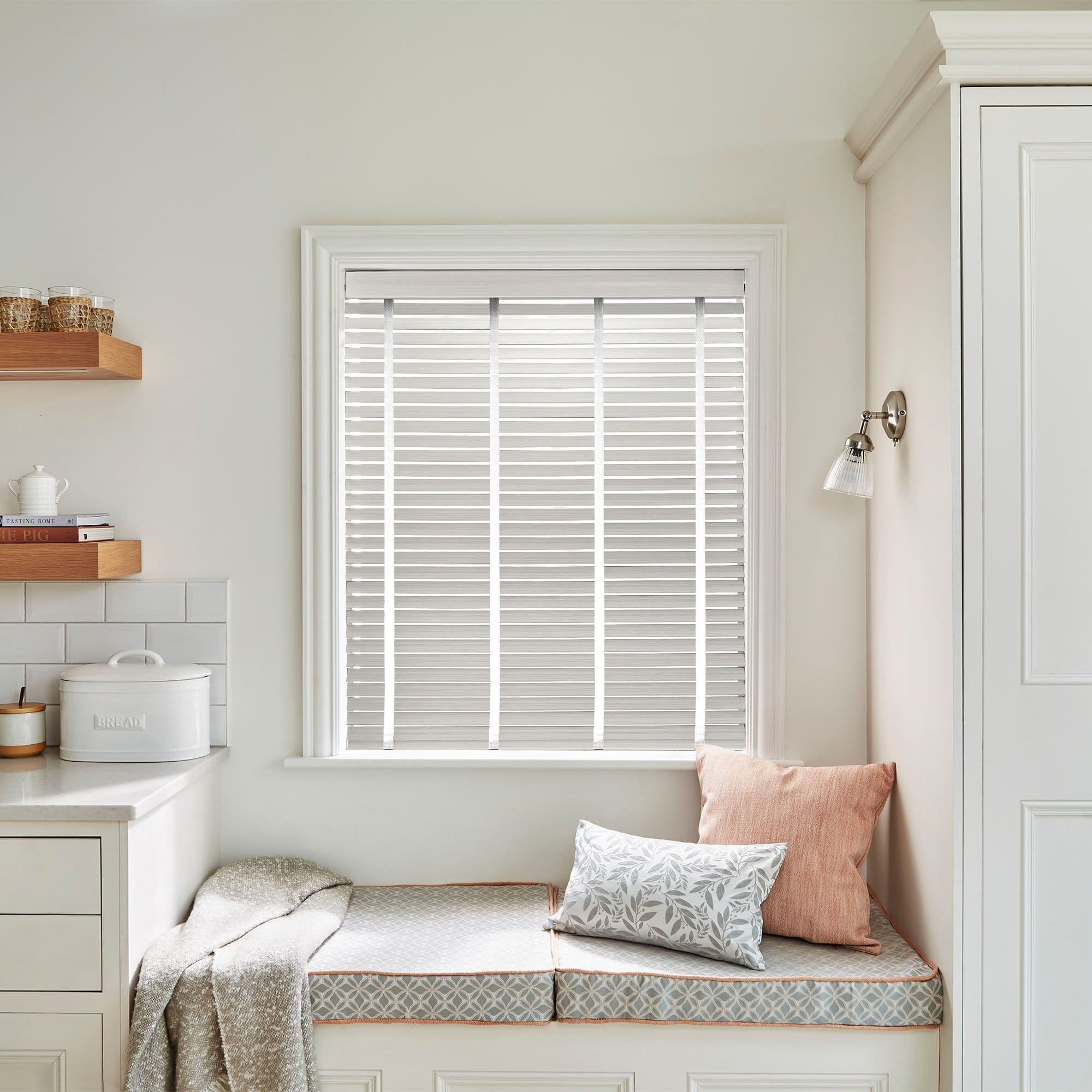 White horizontal Venetian blinds inside the window frame with narrow vertical tapes â€” closed, filtering soft daylight â€” above a cozy kitchen window seat with cushions, pale cabinetry, and a wall sconce.

BREAD