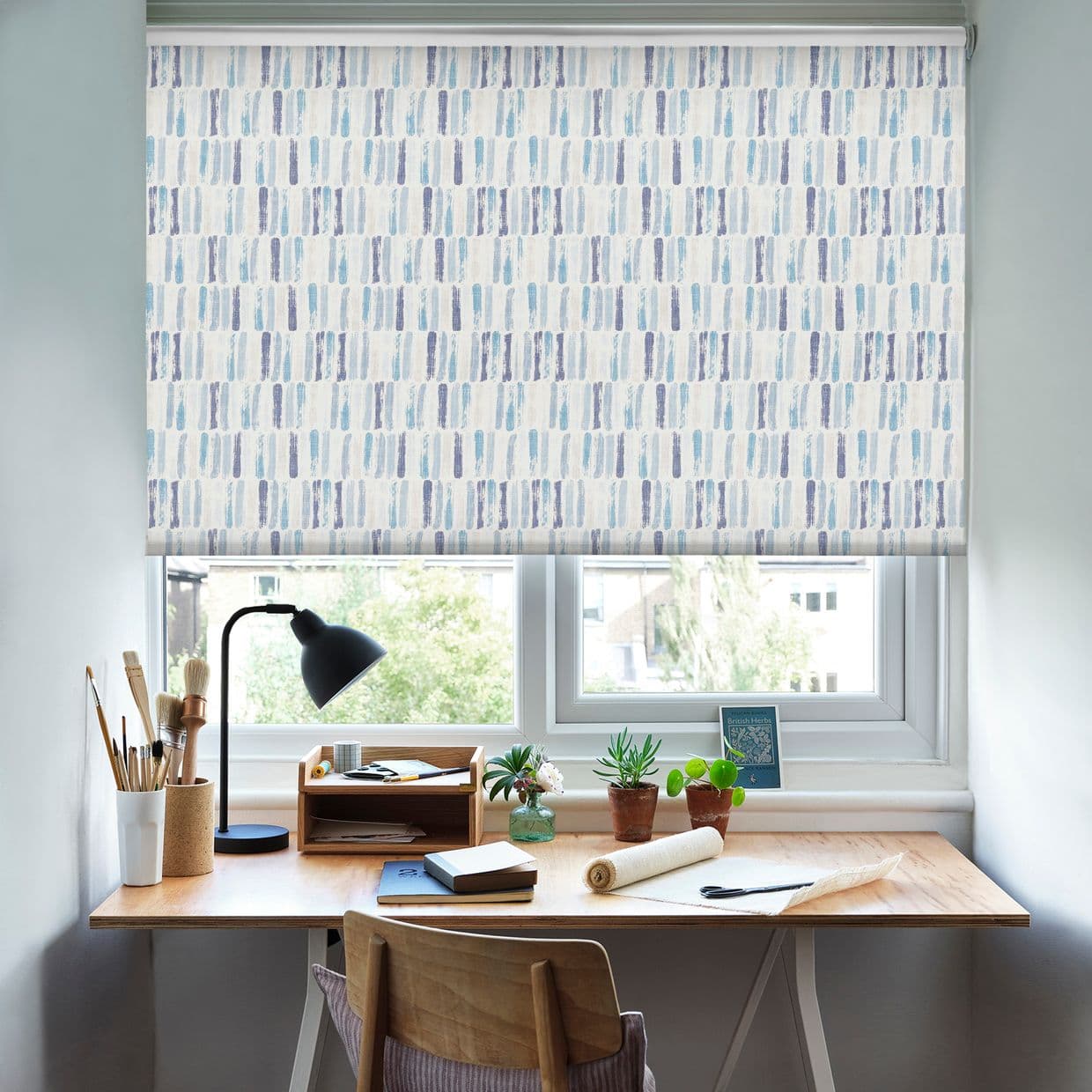 Patterned roller blind â€” white with vertical blue brushstroke motif â€” lowered across a double window, filtering soft daylight; a wooden desk beneath holds a lamp, potted plants, notebooks, and art tools.