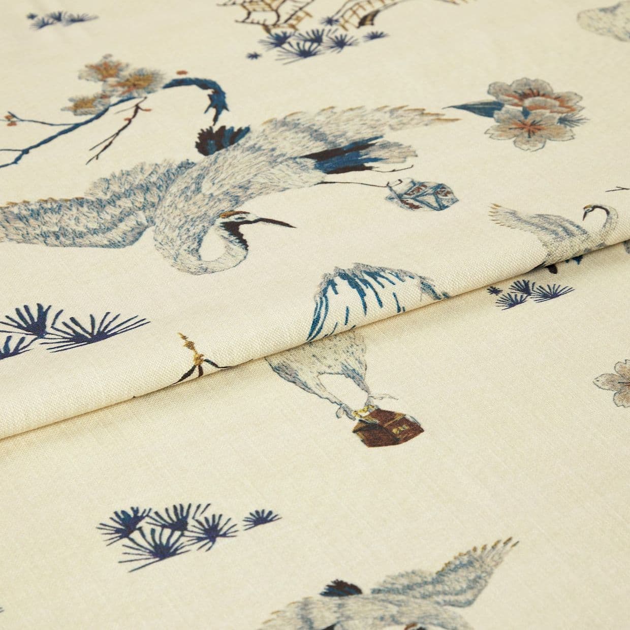 Light cream fabric with blue-grey crane and floral print, folded once; pattern visible across woven texture, photographed in soft studio light as a close-up product shot.