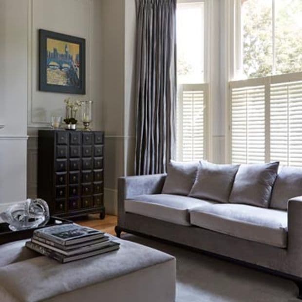 Floor-to-ceiling pleated gray curtains alongside white cafÃ©-style louvered shutters covering a bay window, partially closed and filtering bright daylight; modern living room with gray sofa, ottoman, dark cabinet.