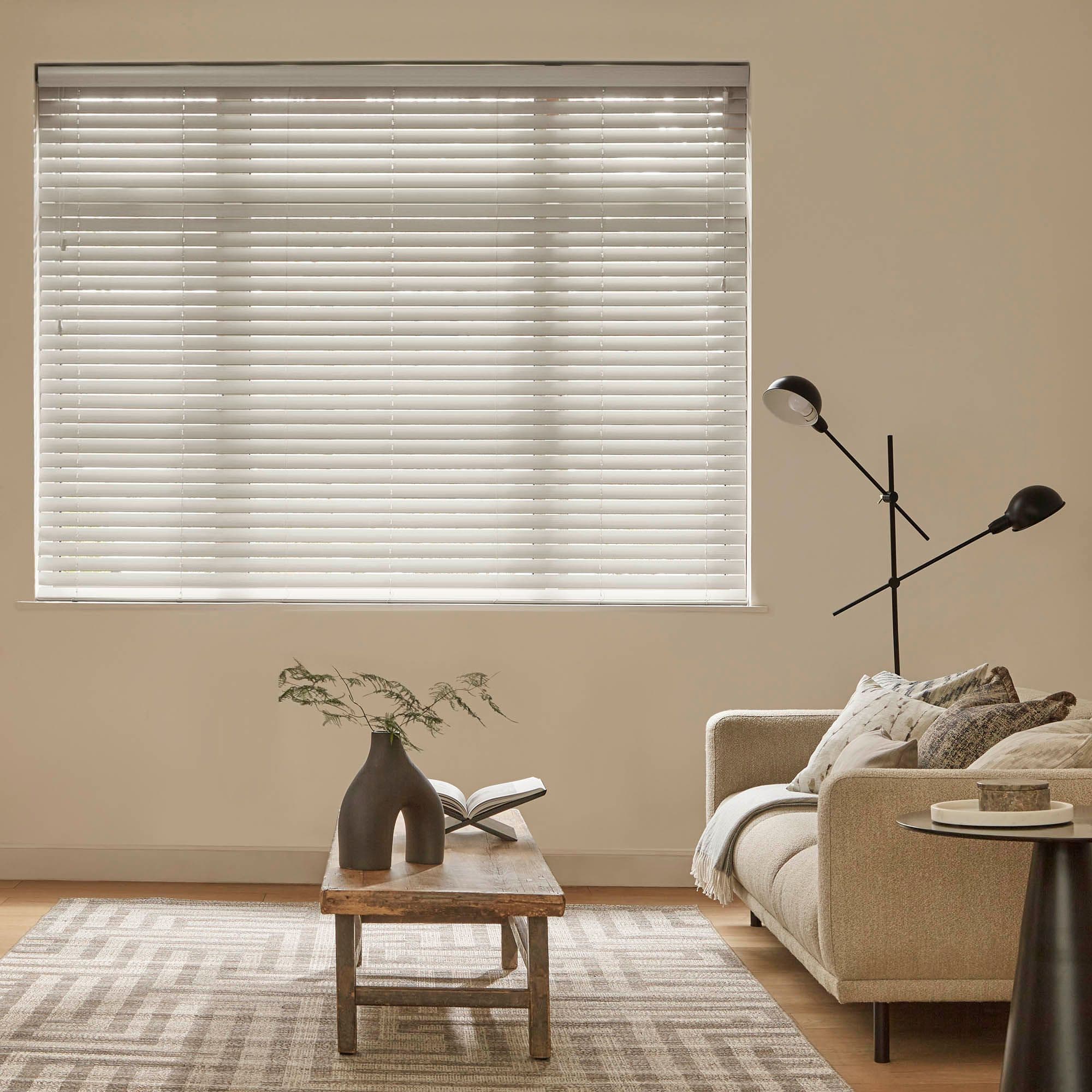 White horizontal venetian blinds cover a large rectangular window; slats tilted closed, filtering soft daylight into a neutral-toned living room with sofa, wooden coffee table, rug, and black floor lamp.