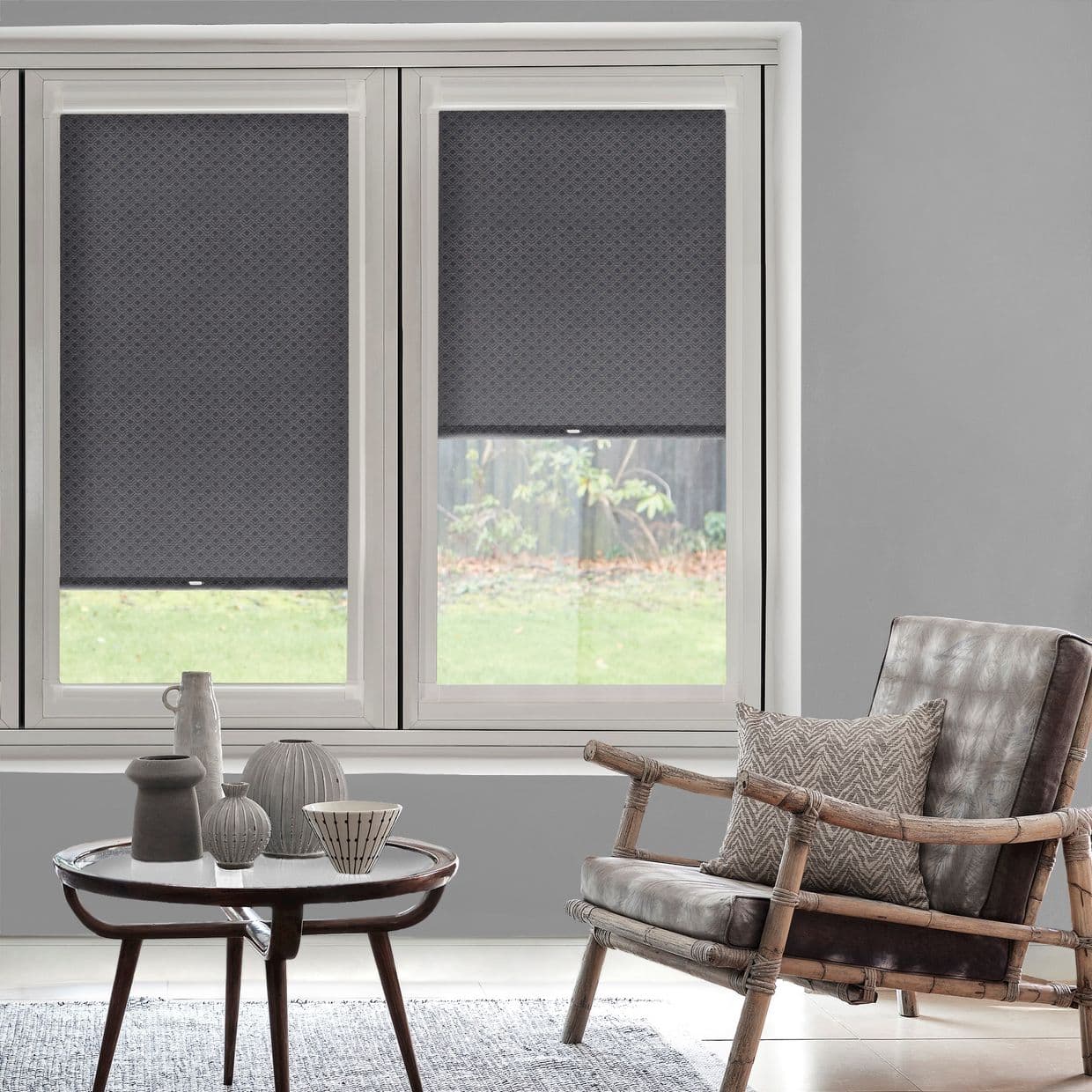 Three grey patterned roller blinds cover a triple window â€” the right blind partially raised, filtering soft daylight into a neutral living room with a rattan armchair and small decorative table.