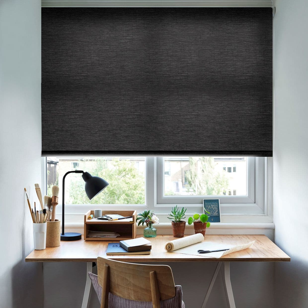 A charcoal textured roller blind, lowered across the window and blocking light; over a sunlit home workspace with a wooden desk, lamp, art supplies, notebooks and small potted plants.