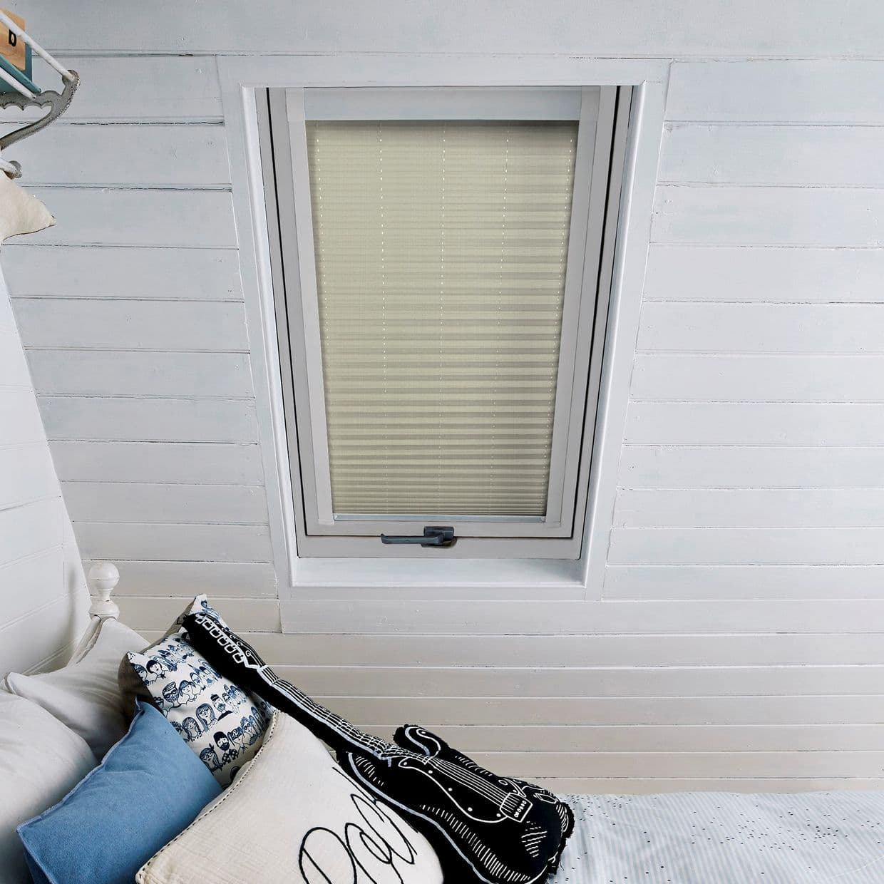 Beige pleated blind, fully lowered in a skylight in an angled white wood-paneled ceiling; filtering soft daylight into an attic bedroom with blue and patterned pillows and a guitar cushion.