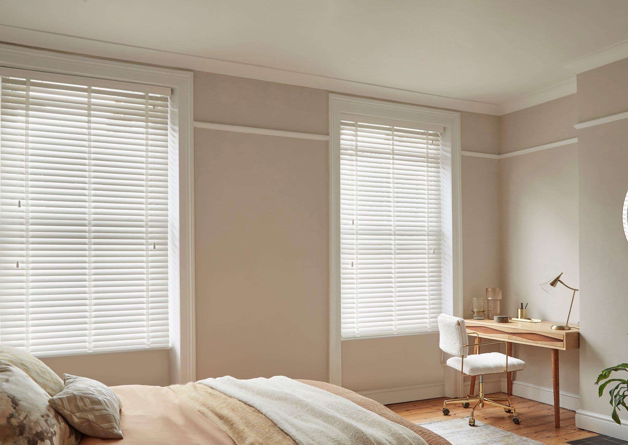 White horizontal blinds covering two tall windows, fully lowered with slats slightly tilted, filtering soft daylight into a neutral-toned bedroom with bed, wooden desk, upholstered chair and warm natural light.