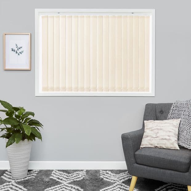 Cream vertical blinds covering a rectangular window, slats closed and gently diffusing daylight; set in a gray-walled living room with a potted plant, framed botanical print, gray armchair, patterned rug.