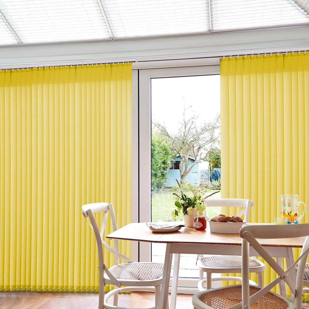 Yellow vertical blinds across patio doors, mostly closed with a central opening, filtering daylight; small dining table and chairs in foreground, sunlit room overlooking garden with tree and shed.