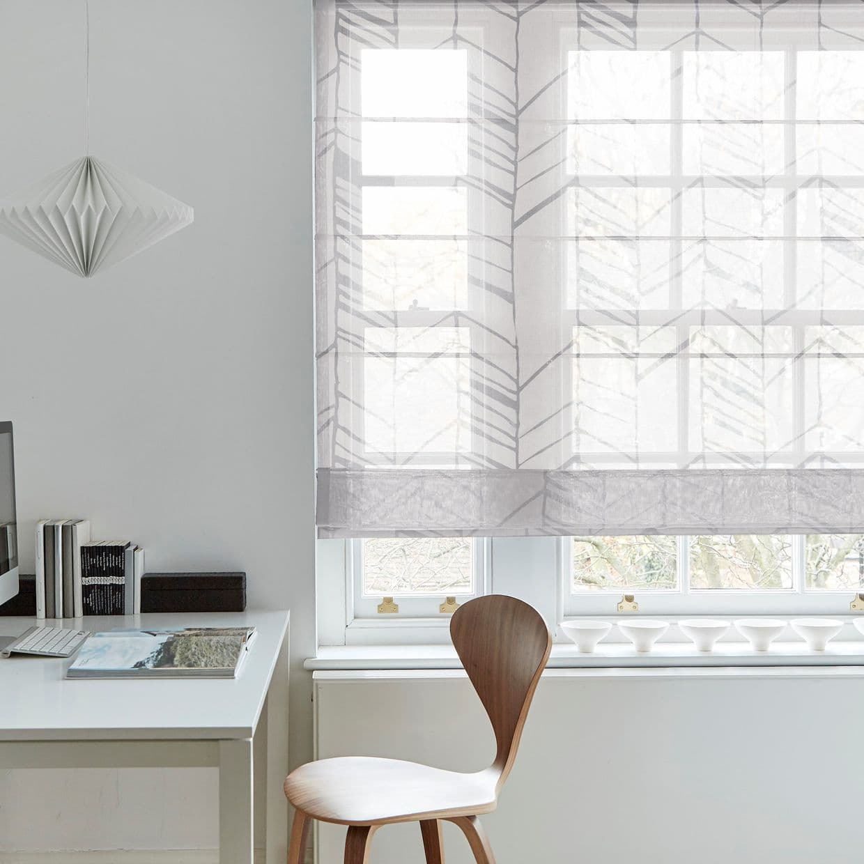 Sheer patterned Roman blind lowered over double sash windows, semi-transparent chevron leaf design filtering soft daylight; in a bright minimalist home office with a white desk, wooden chair, pendant lamp.