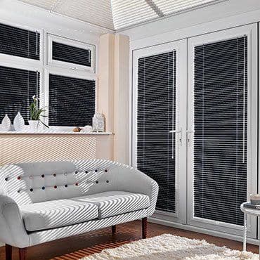 White internal Venetian blinds on a bay window and glazed French doors, closed with narrow slats slicing sunlight into sharp stripes, in a bright living room with pale upholstered sofa, floor.
