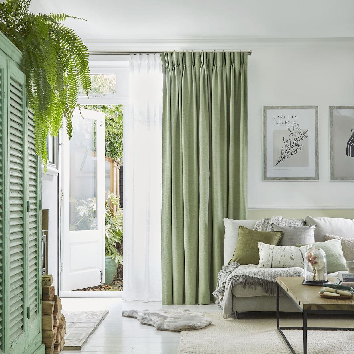 Green pinch-pleated curtains with an inner white sheer, partly drawn and filtering daylight beside an open white door. Bright, plant-filled living room with pale sofa, cushions, wooden coffee table, botanical wall art.

L'ART DES FLEURS