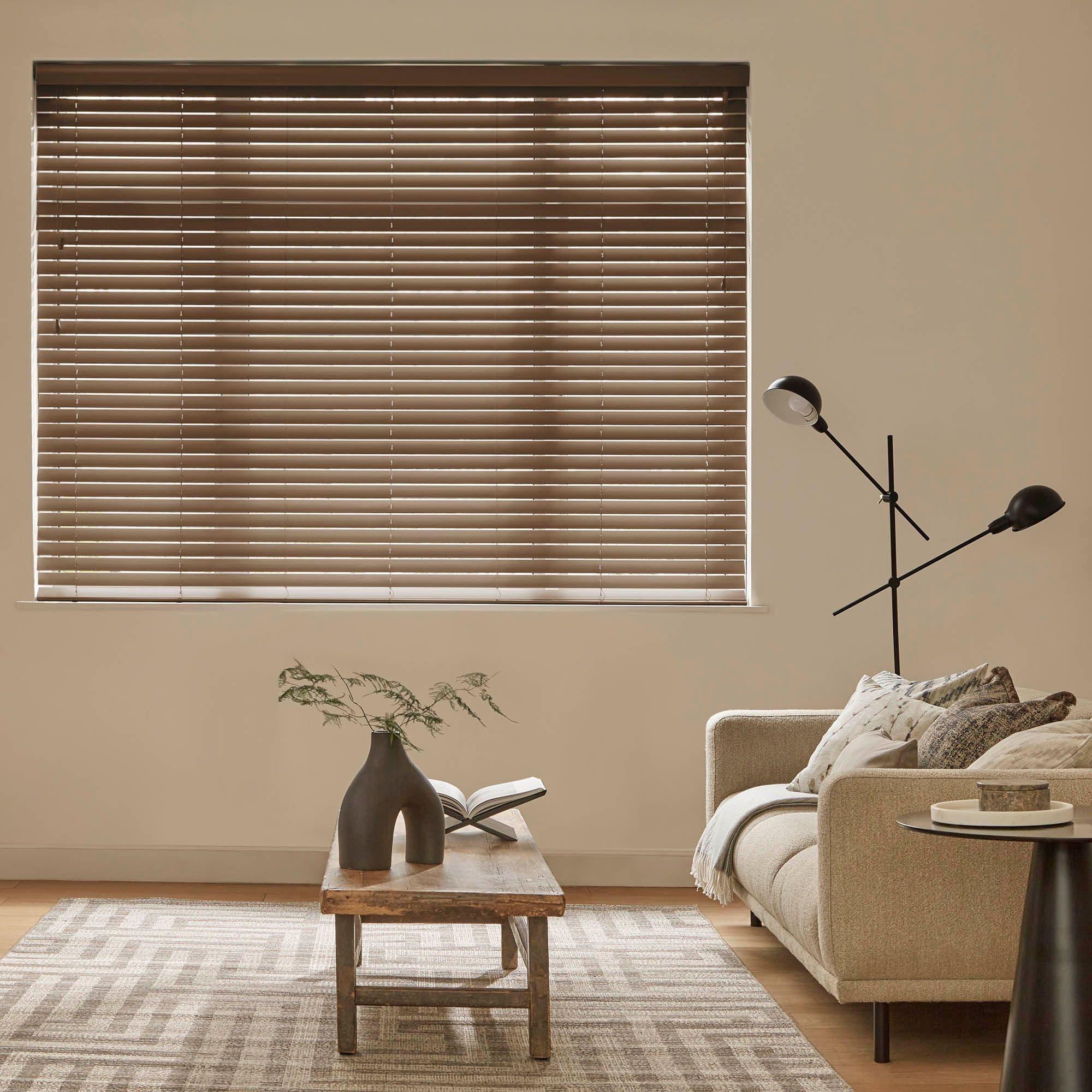 Horizontal wooden Venetian blinds covering a window, fully closed and filtering daylight through narrow slats; beige living room with sofa, wooden coffee table, sculptural vase, open book and floor lamp.