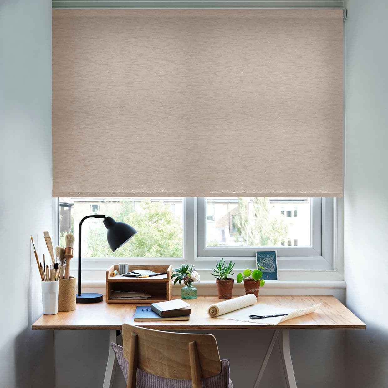Beige roller blind lowered across the window, textured fabric filtering daylight; wooden desk with lamp, brushes, notebooks and potted plants beneath; small home office overlooking a leafy suburban view.