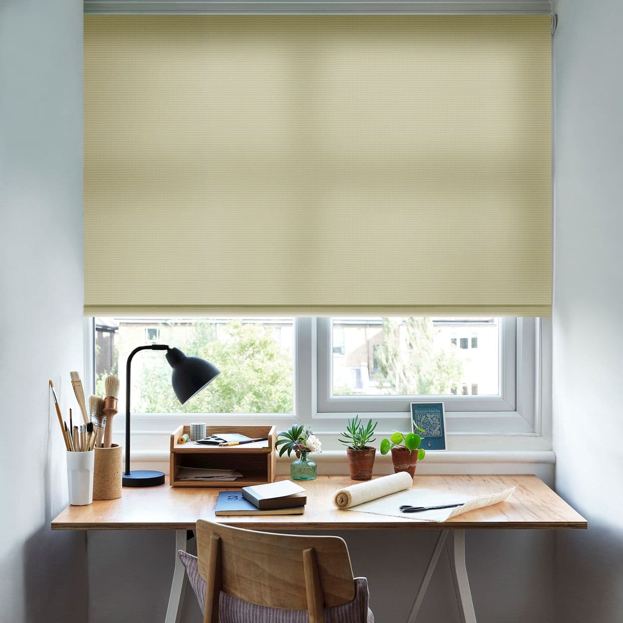 Beige fabric roller blind, lowered almost fully across the window with subtle grid texture; filtering daylight; home workspace below with wooden desk, black lamp, potted plants, notebooks, and chair.