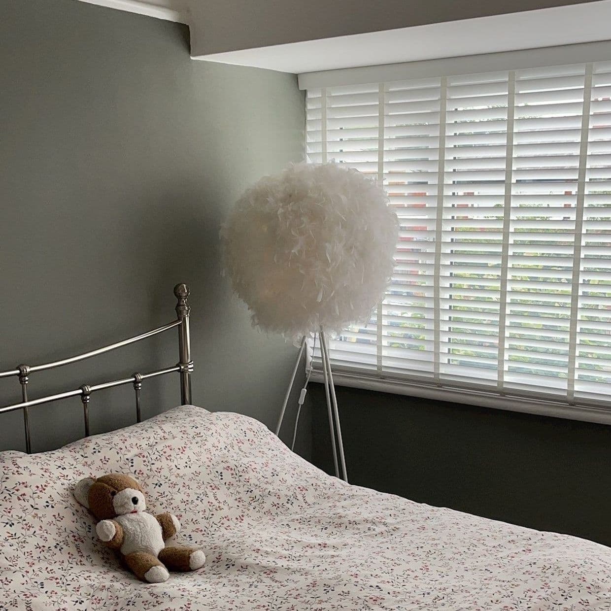 White horizontal slatted blinds cover the right window, filtering soft daylight. Bedroom with muted grey-green walls, metal bedframe, floral duvet, small teddy on bed and a white feathered tripod lamp.