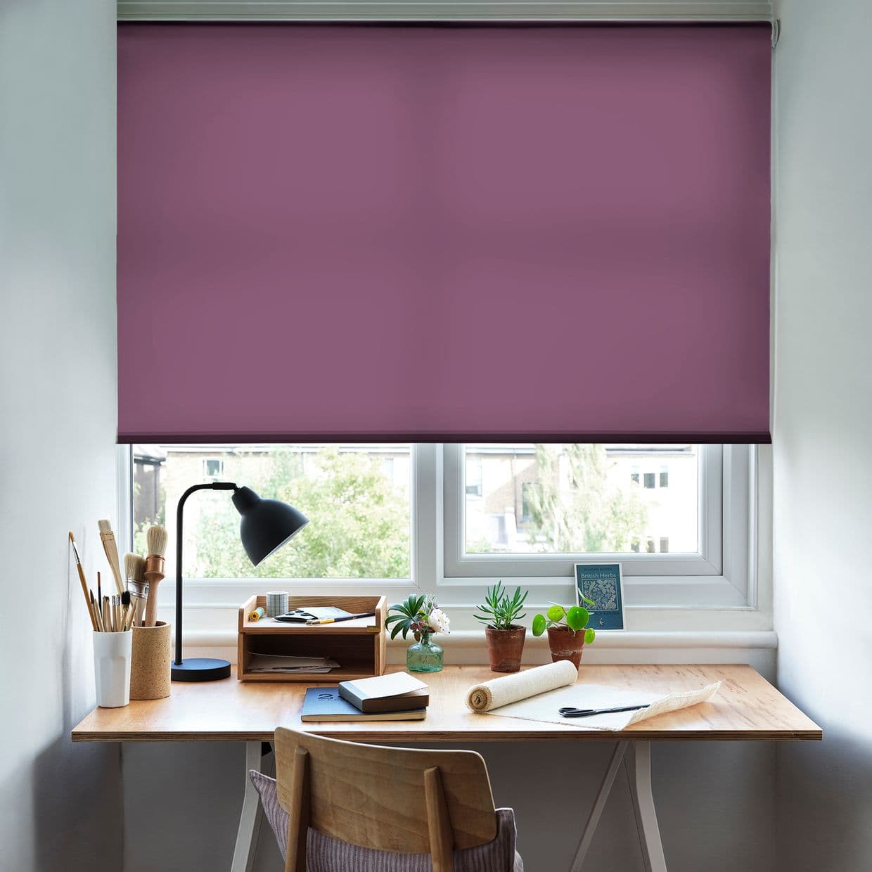 Plum-purple roller blind, lowered nearly fully closed with a narrow bottom gap; filtering soft daylight; small home workspace with a wooden desk, lamp, brushes, notebooks and potted plants.