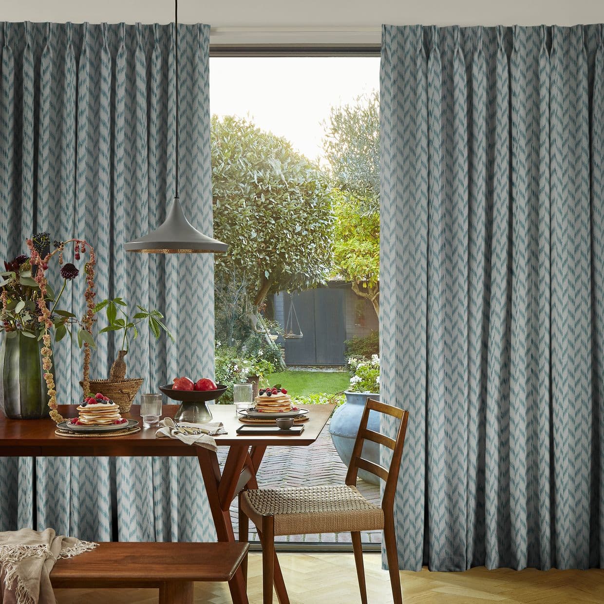 Floor-to-ceiling pleated blue-gray curtains with a chevron-like pattern, parted at center; filtering soft morning light. Dining table with plates and pendant lamp in foreground, green garden visible through glass door.