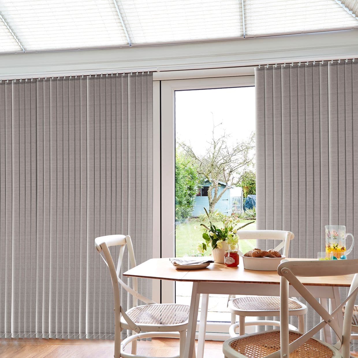 Grey fabric vertical blinds, hung across a sliding glass door, partly drawn aside to reveal a sunlit backyard; breakfast table and white chairs sit in a bright dining nook.