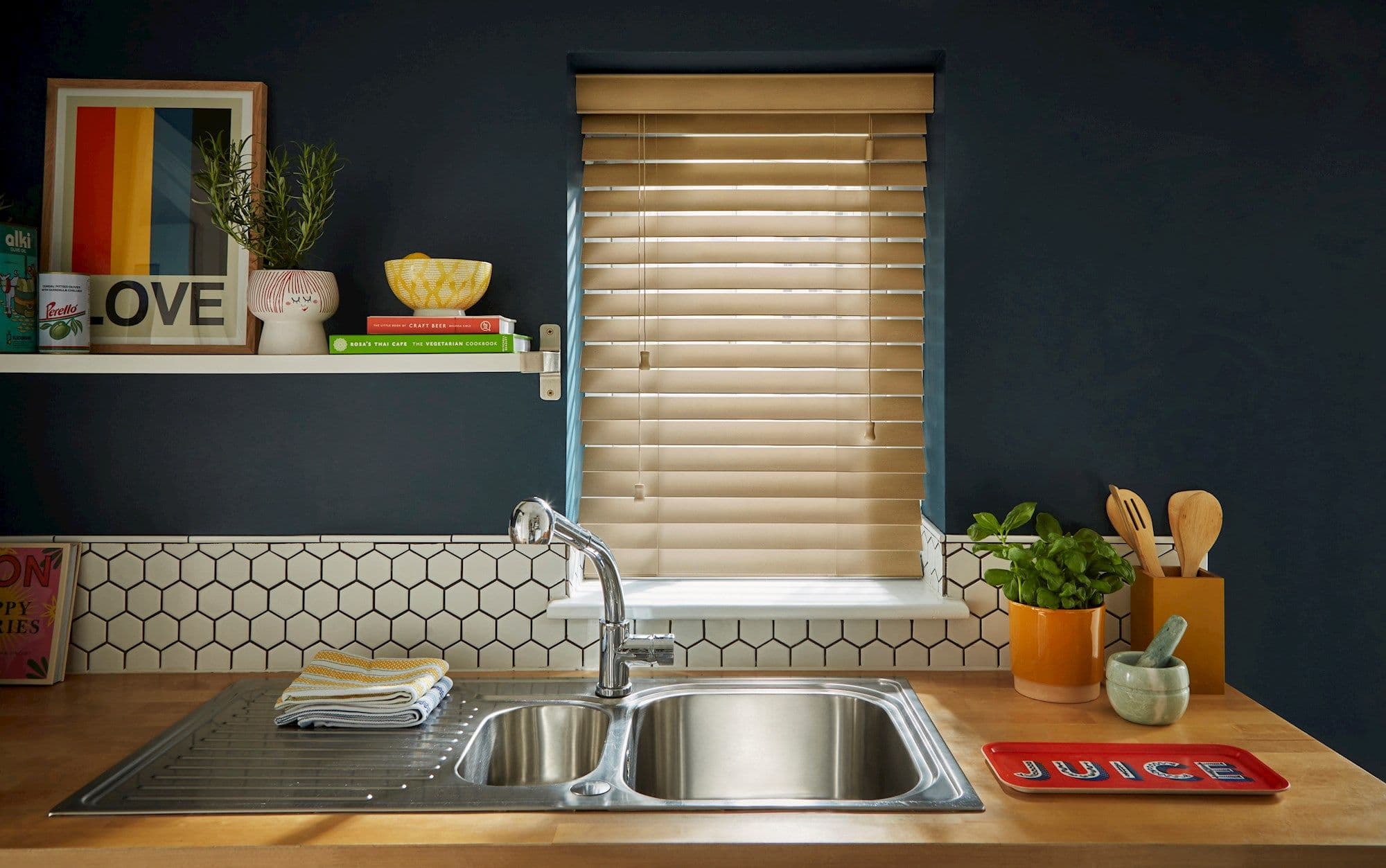 Beige wooden Venetian blinds centered in frame, closed and filtering warm daylight through slats; above a stainless double sink in a dark-blue kitchen with hex-tile backsplash, shelf, plants, utensils.

LOVE
JUICE