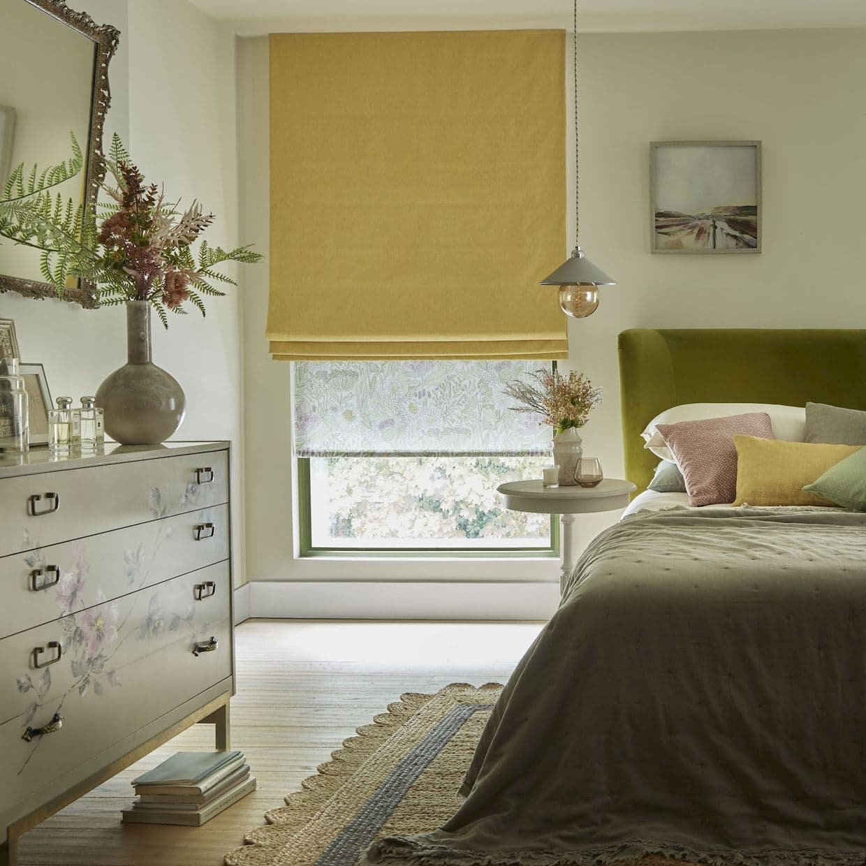 Yellow Roman blind lowered over a window with a patterned sheer beneath, filtering daylight into a cozy bedroom with an oliveâ€‘green upholstered headboard, bedside table, floralâ€‘painted dresser, rug, and pendant light.