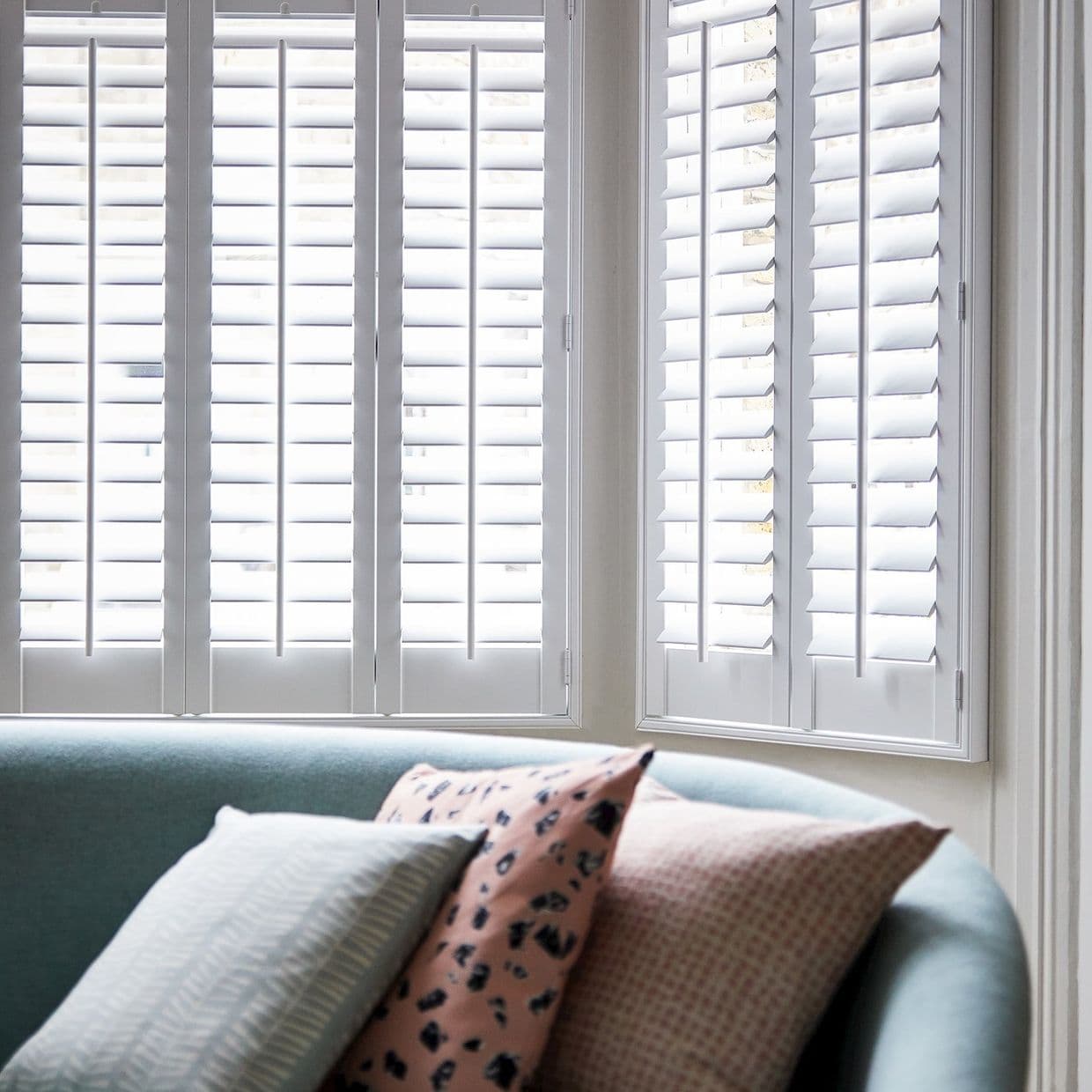 White plantation shutters covering corner bay windows, louvered panels tilted to diffuse bright daylight; in front, a curved teal sofa with patterned cushions in a softly lit living room.