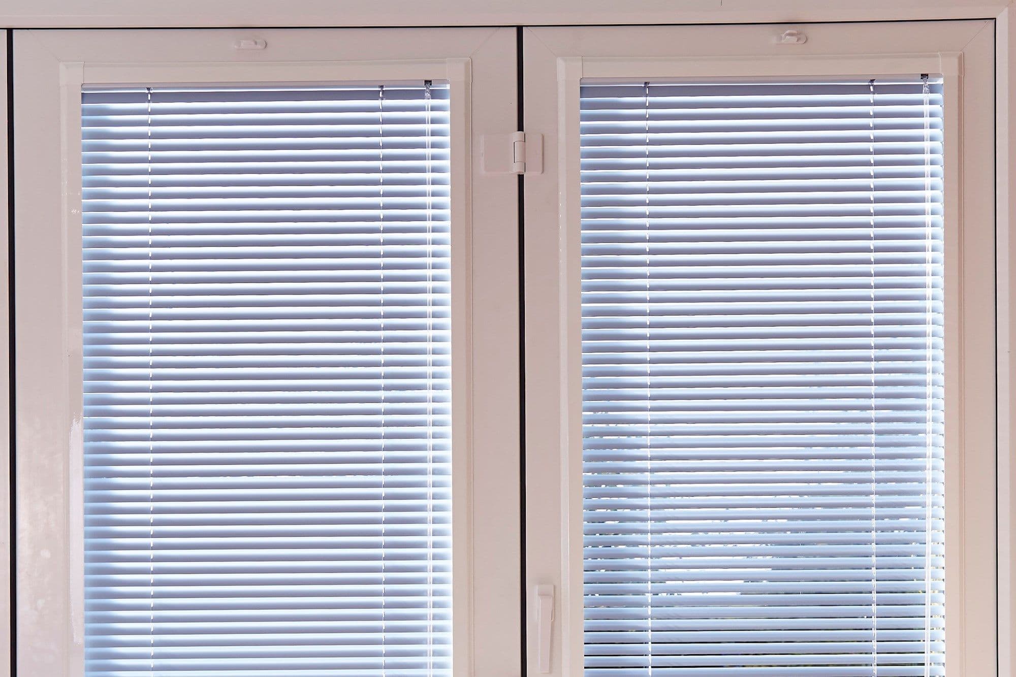 White horizontal Venetian blinds covering a pair of narrow glass panels; slats closed, filtering daylight into thin horizontal bands; set within white double-door frames with visible handle and hinge.