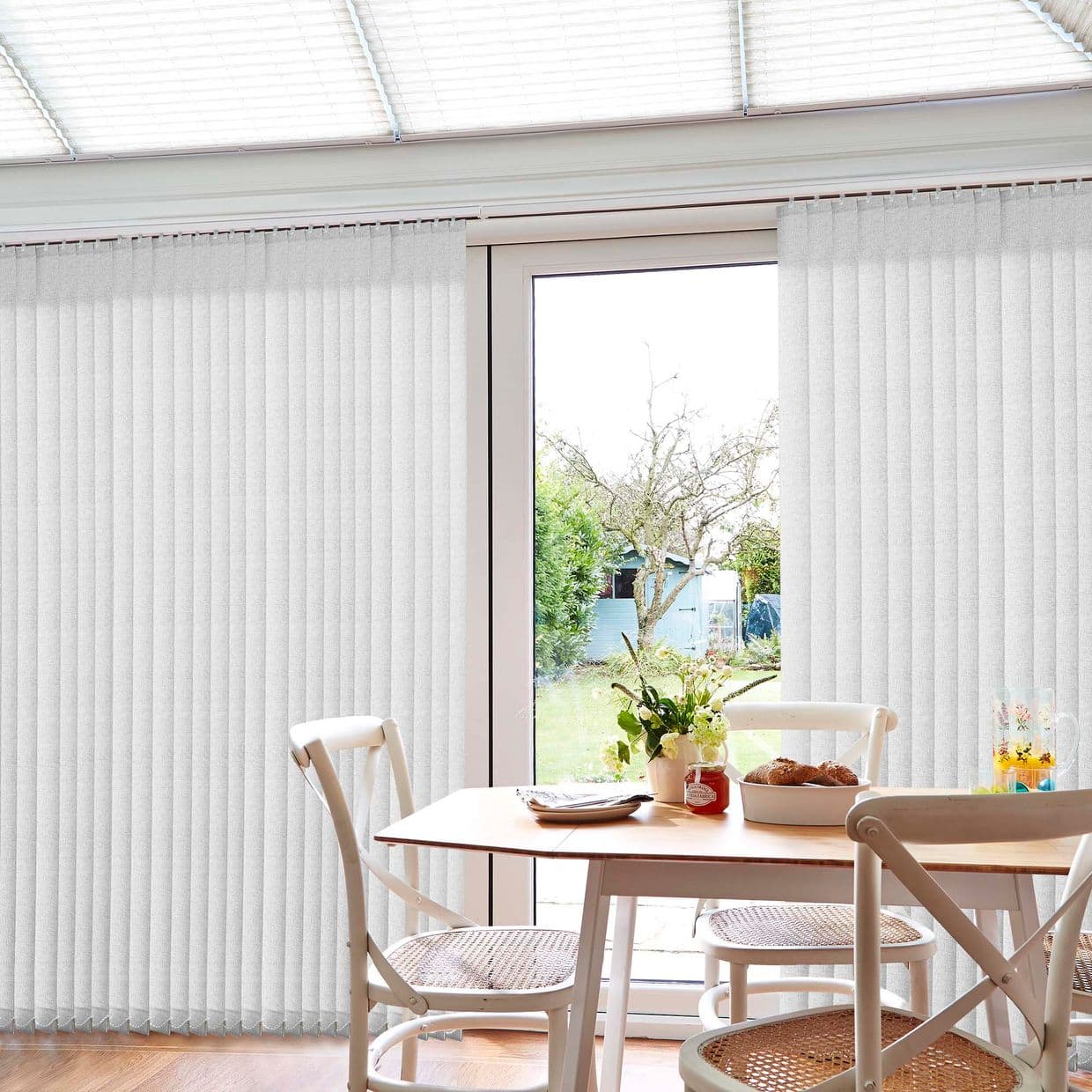 Vertical white blinds on a track, mostly closed but parted to reveal a glass patio door; filtering daylight into a dining area with wooden table and chairs, garden beyond.