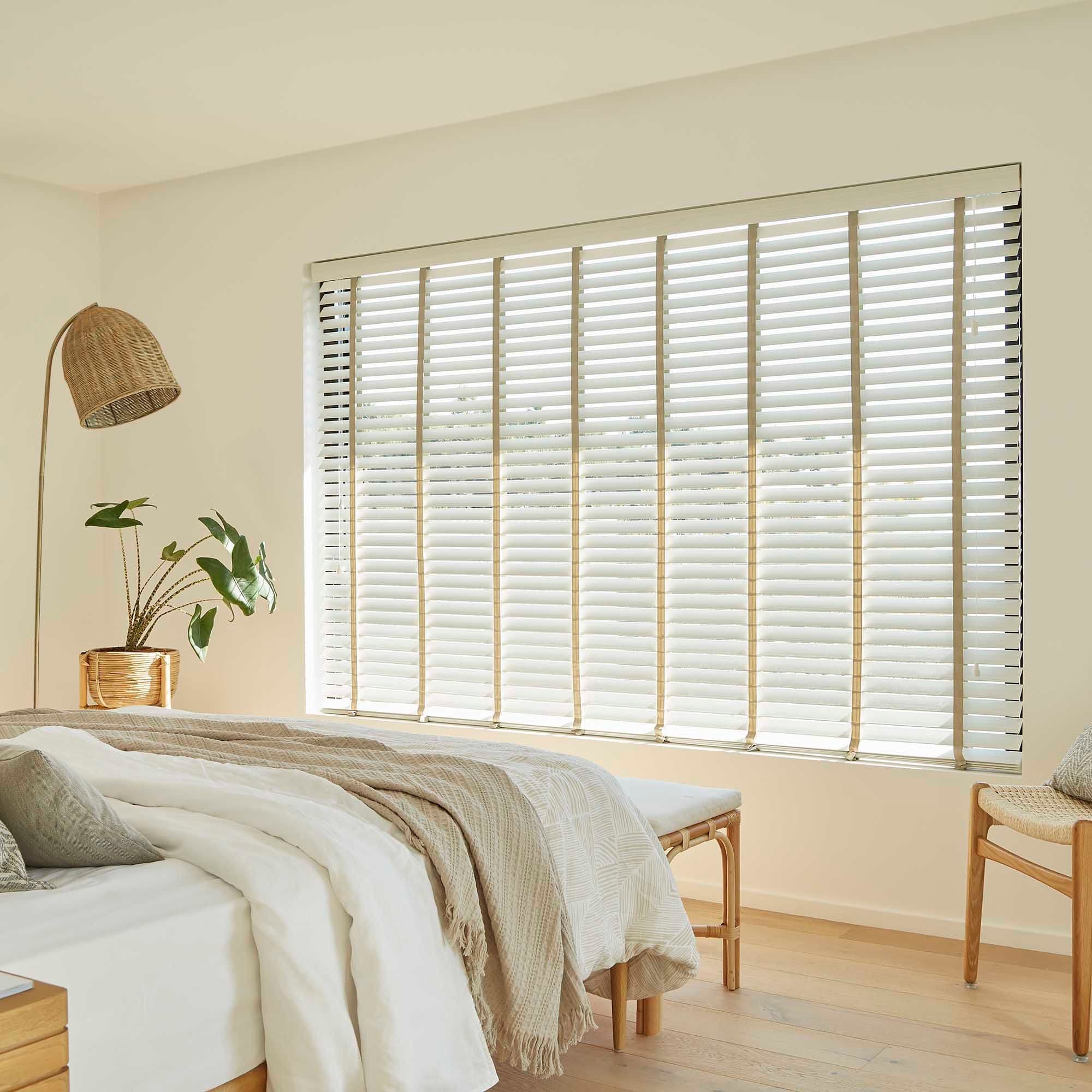 Horizontal wood slat blinds cover a large window, slats tilted to diffuse daylight; in a minimalist sunlit bedroom with neutral linens, a rattan lamp, potted plant, and light wood floors.