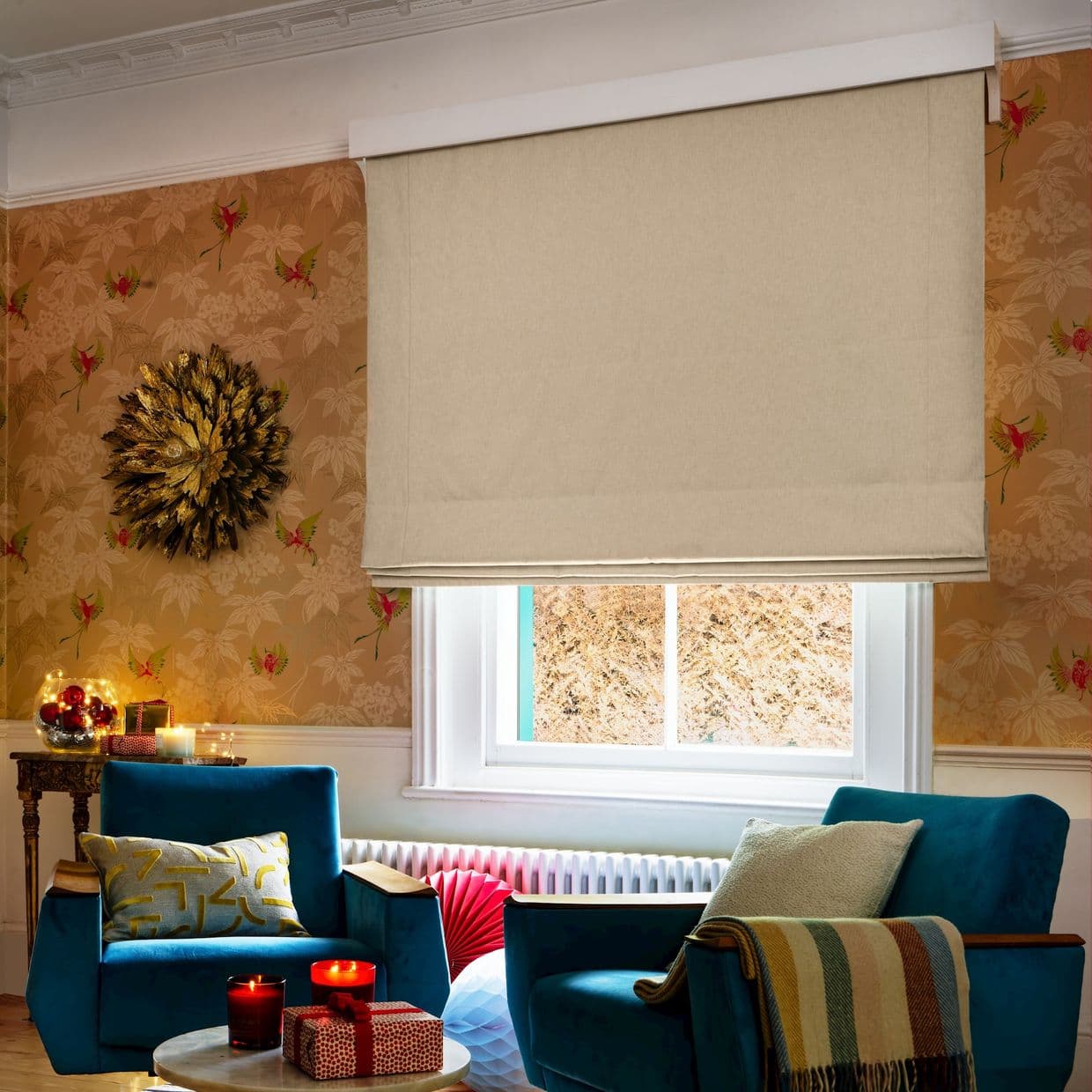 A beige fabric Roman blind, lowered with soft horizontal folds, covers a white-framed window, softly diffusing daylight in a cozy living room with teal armchairs, patterned wallpaper, warm accent lighting.