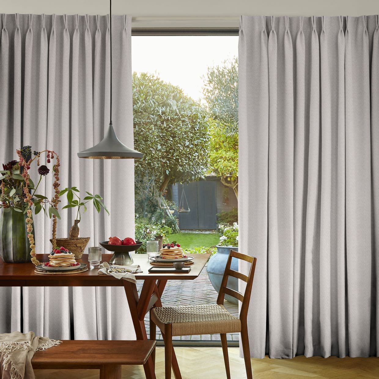 Pleated light-gray curtains, full-length and hung high, parted centrallyâ€”partially drawn and filtering daylightâ€”framing a breakfast dining table with pancakes and a sunlit garden beyond with trees and a shed.