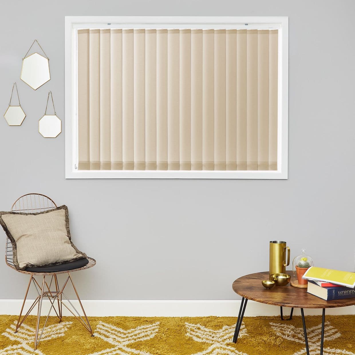 Beige vertical blinds on a white-framed window, closed and filtering light; grey-walled living room with wire chair with cushion, round wooden coffee table, mustard rug, three hanging hexagonal mirrors.