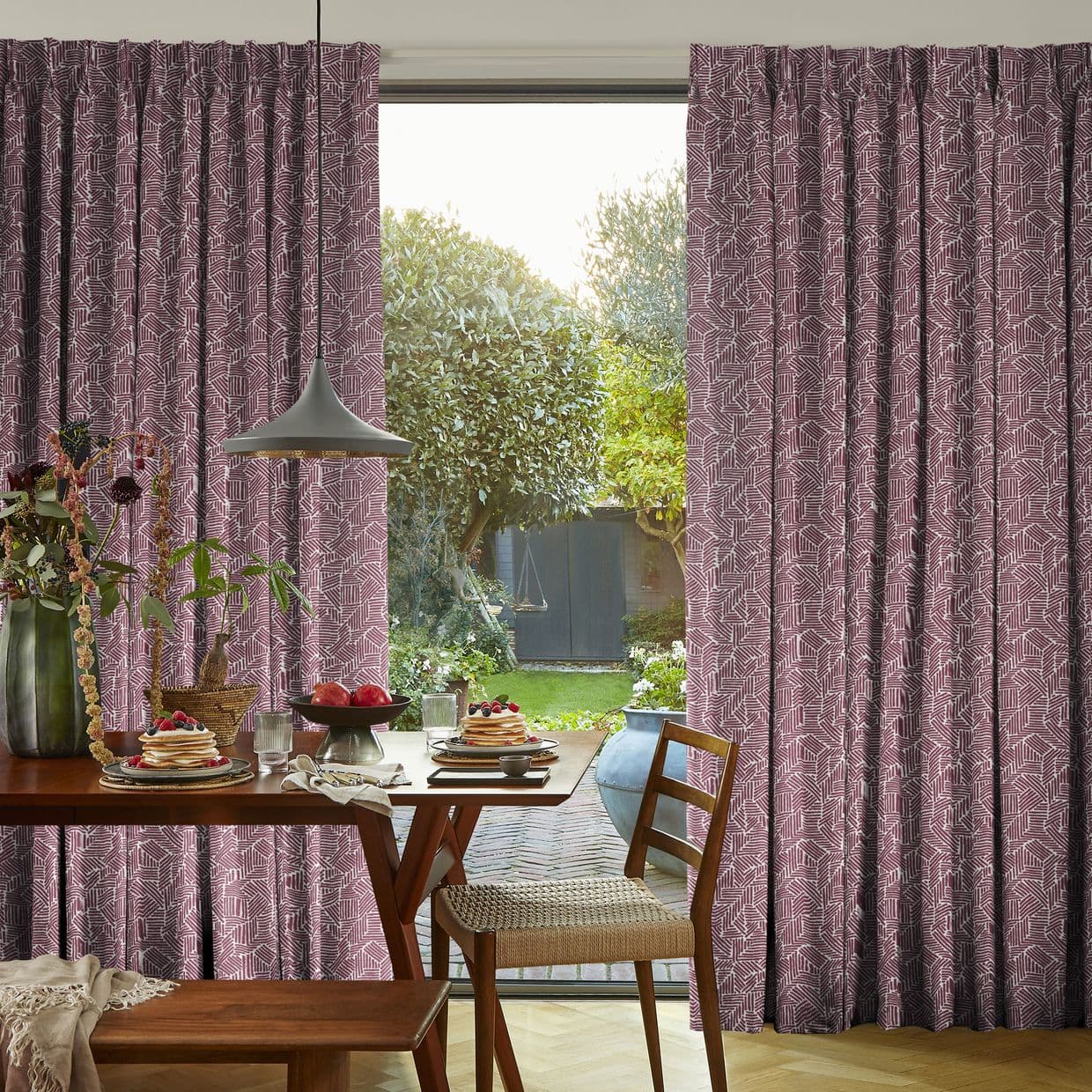 Pleated mauve curtains with geometric print, floor-length and drawn aside, parting a sliding door to frame a sunlit backyard. Wooden dining table set for breakfast beneath a pendant lamp.