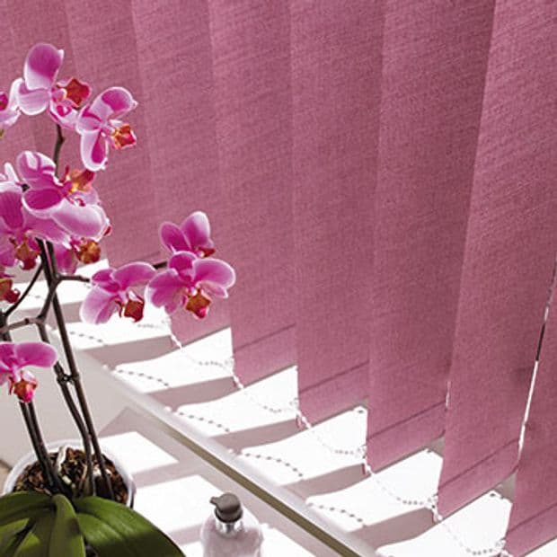 Vertical fabric blinds in dusty rose cover a window, slightly overlapping and filtering bright sunlight into angled bands; on the windowsill a potted pink orchid and bottle catch the light.