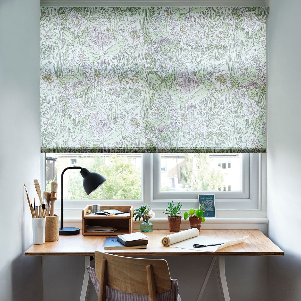 A floral-patterned roller blind, fully lowered and filtering soft daylight; covers a double window above a wooden desk with lamp, paintbrushes, small potted plants, and stationery in a bright study.