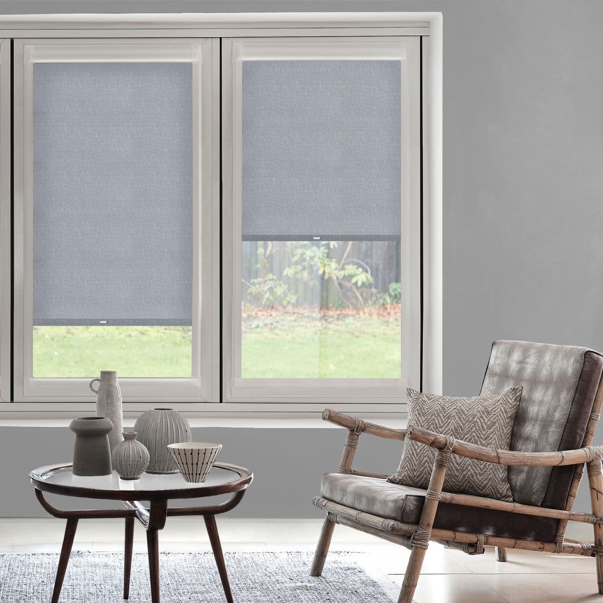 Two light-gray fabric roller blinds covering a double window, partly lowered and filtering daylight; modern living room with neutral walls, rattan armchair with cushion, round coffee table and garden view.
