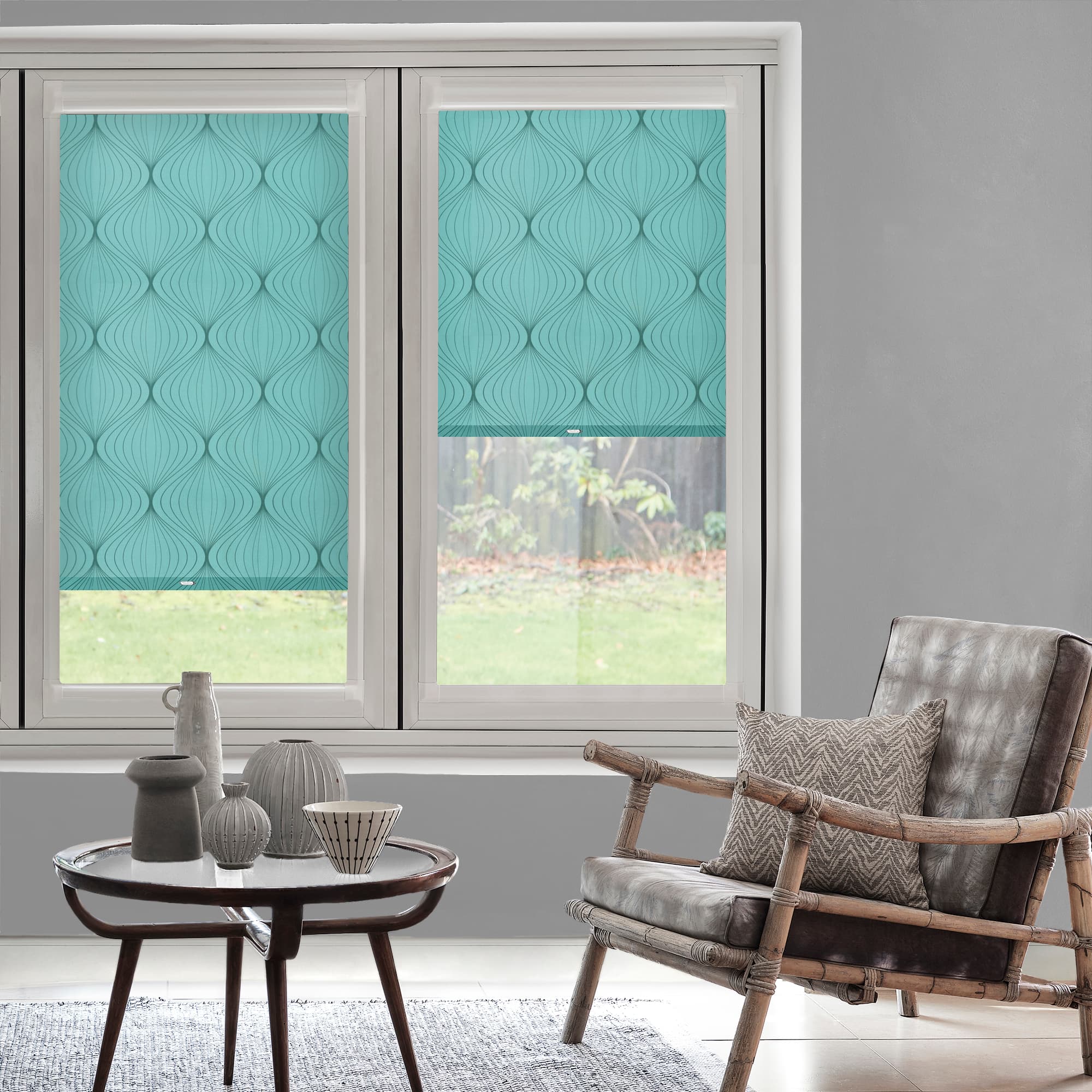 Turquoise, leaf-patterned roller blinds covering two sash windowsâ€”one fully lowered, one halfwayâ€”filtering soft daylight; living-room setting with gray walls, rattan armchair, patterned cushion, wooden coffee table and ceramics, garden view.