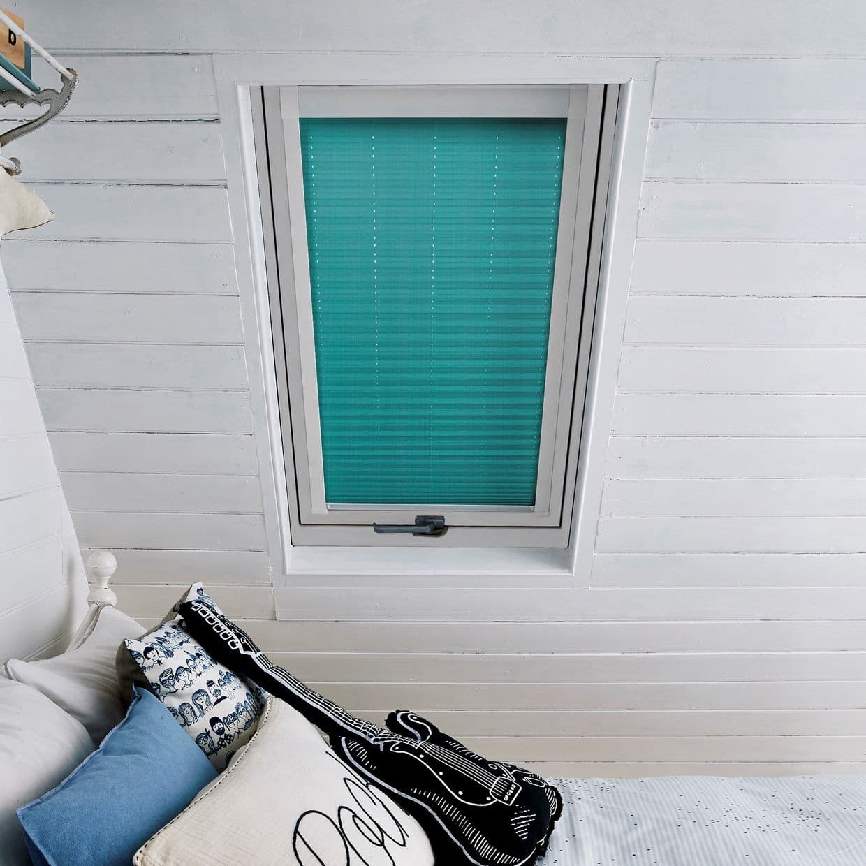 Turquoise pleated blind covers a rectangular skylight, closed and filtering soft daylight; mounted in white painted shiplap ceiling above a bed with blue pillows and a guitar-shaped cushion.