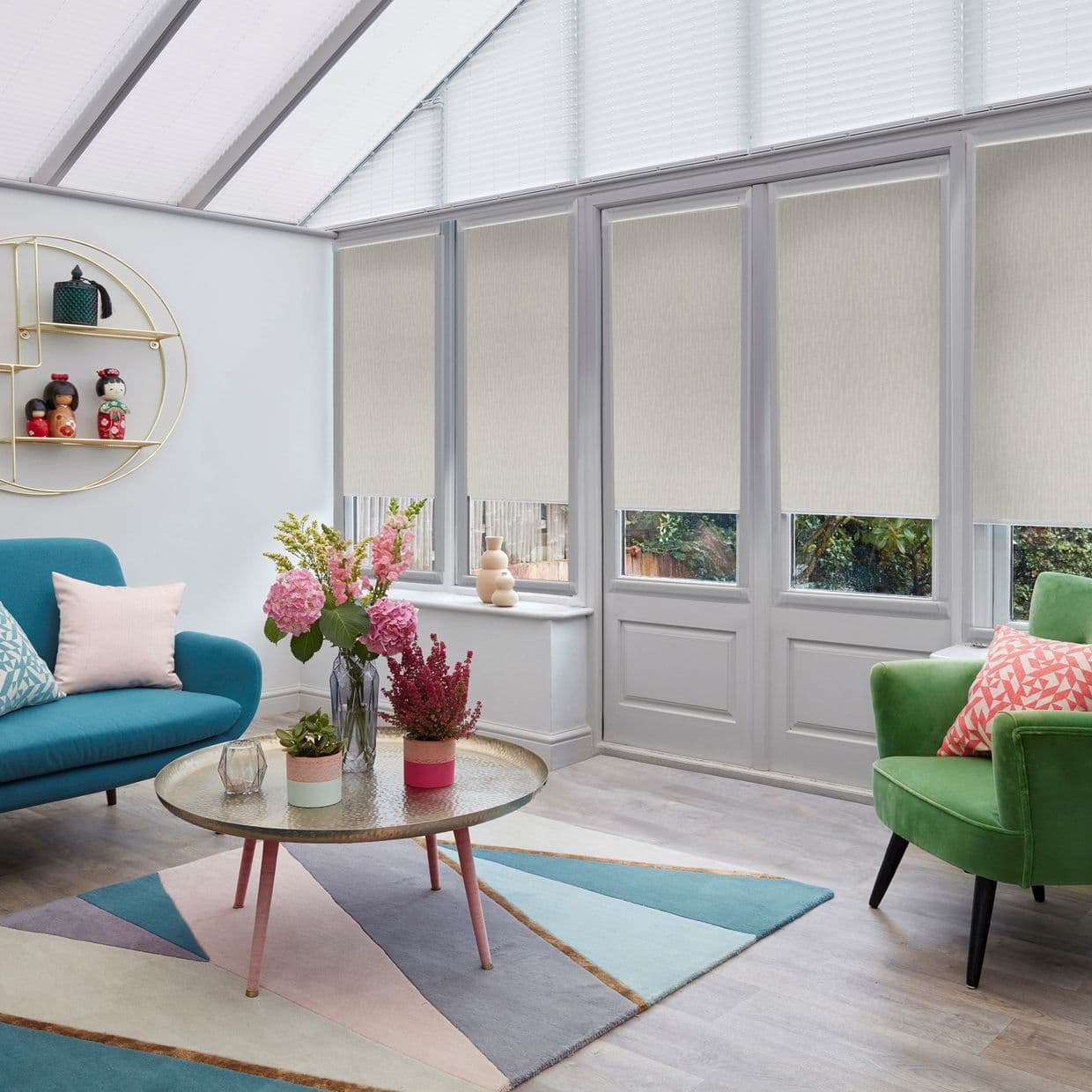 Beige roller blinds cover tall glass doors and windows, lowered halfway and softly filtering daylight; set in a bright conservatory with sloped skylights, teal sofa, green armchair, round coffee table.