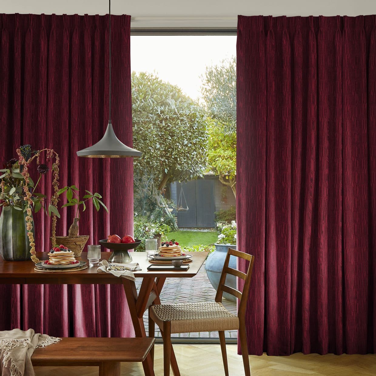 Burgundy pleated curtains, drawn aside to reveal a sunlit garden; a pendant lamp hangs over a dining table set with pancakes and fruit, wooden chairs nearby.