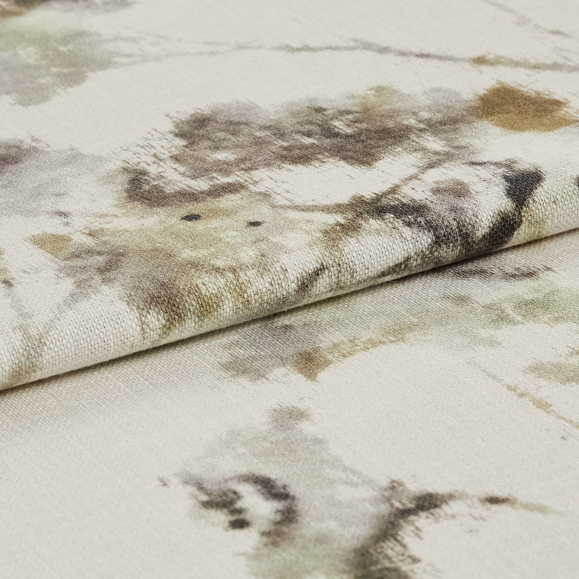 A folded cream fabric with watercolor-style olive, brown, and gray splotches; the folded edge lies across another layer, revealing a textured weave in a lit close-up on a flat surface.