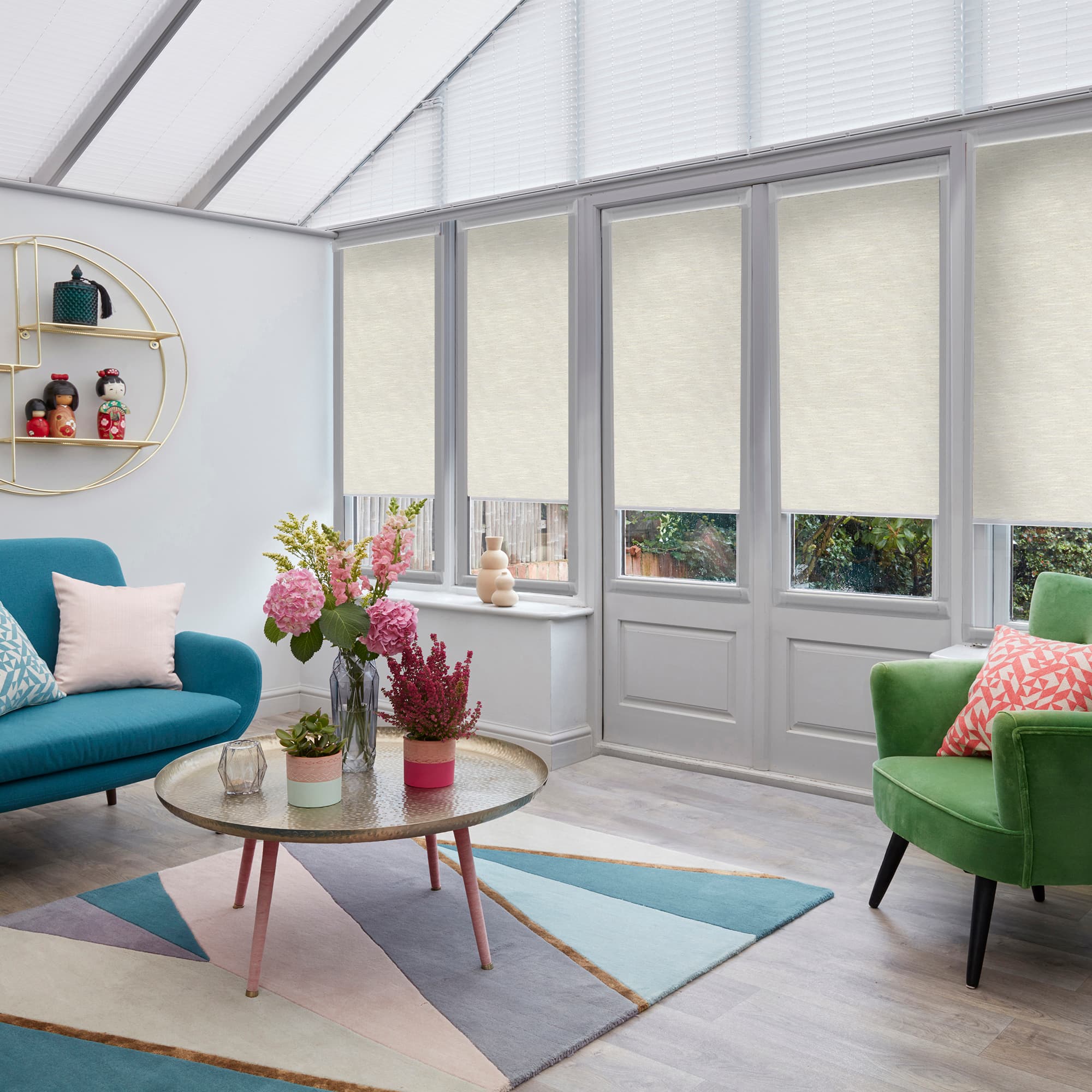Beige roller blinds and pleated roof blinds: lowered across tall conservatory windows and doors, filtering daylight; sunroom with teal sofa, green armchair, round coffee table and floral centerpiece, garden view.