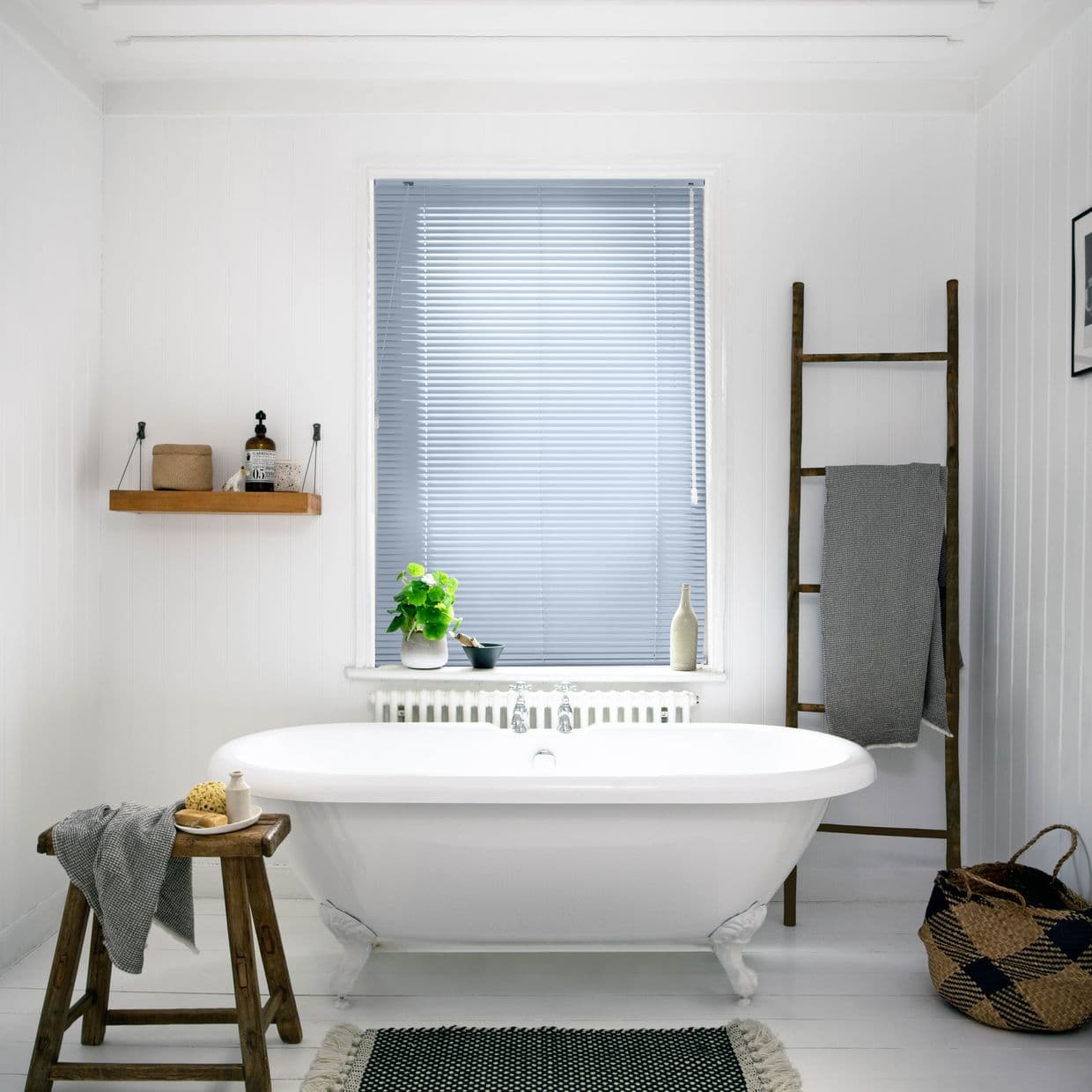 White horizontal venetian blinds covering central window, fully closed and filtering daylight; freestanding clawfoot bathtub beneath, paneled bathroom with wooden stool, ladder towel rack, potted plant on sill.