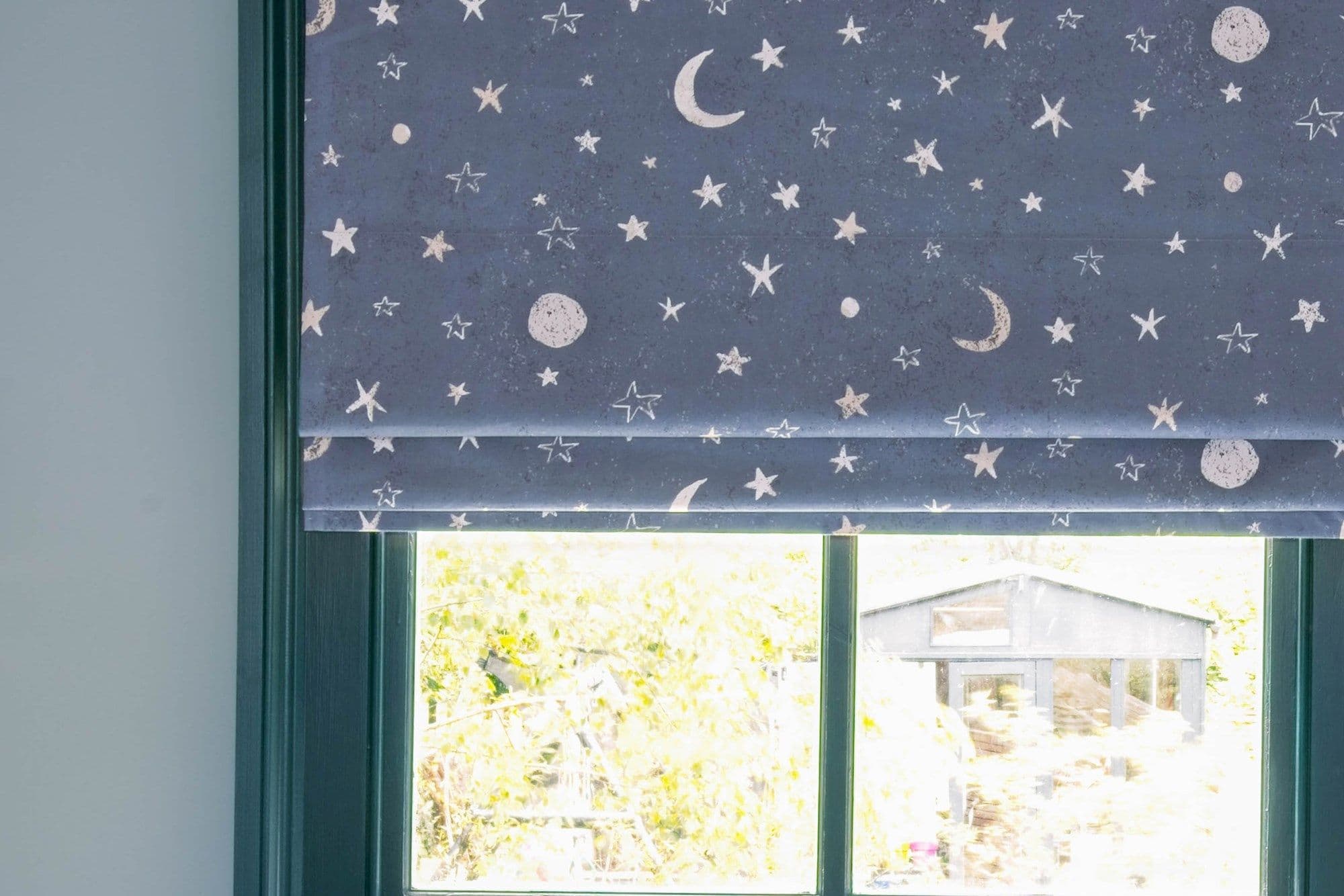 Folded Roman blind with navy fabric patterned with white stars and moons, lowered and filtering daylight; teal window frame reveals a bright sunlit garden and a small shed outside.