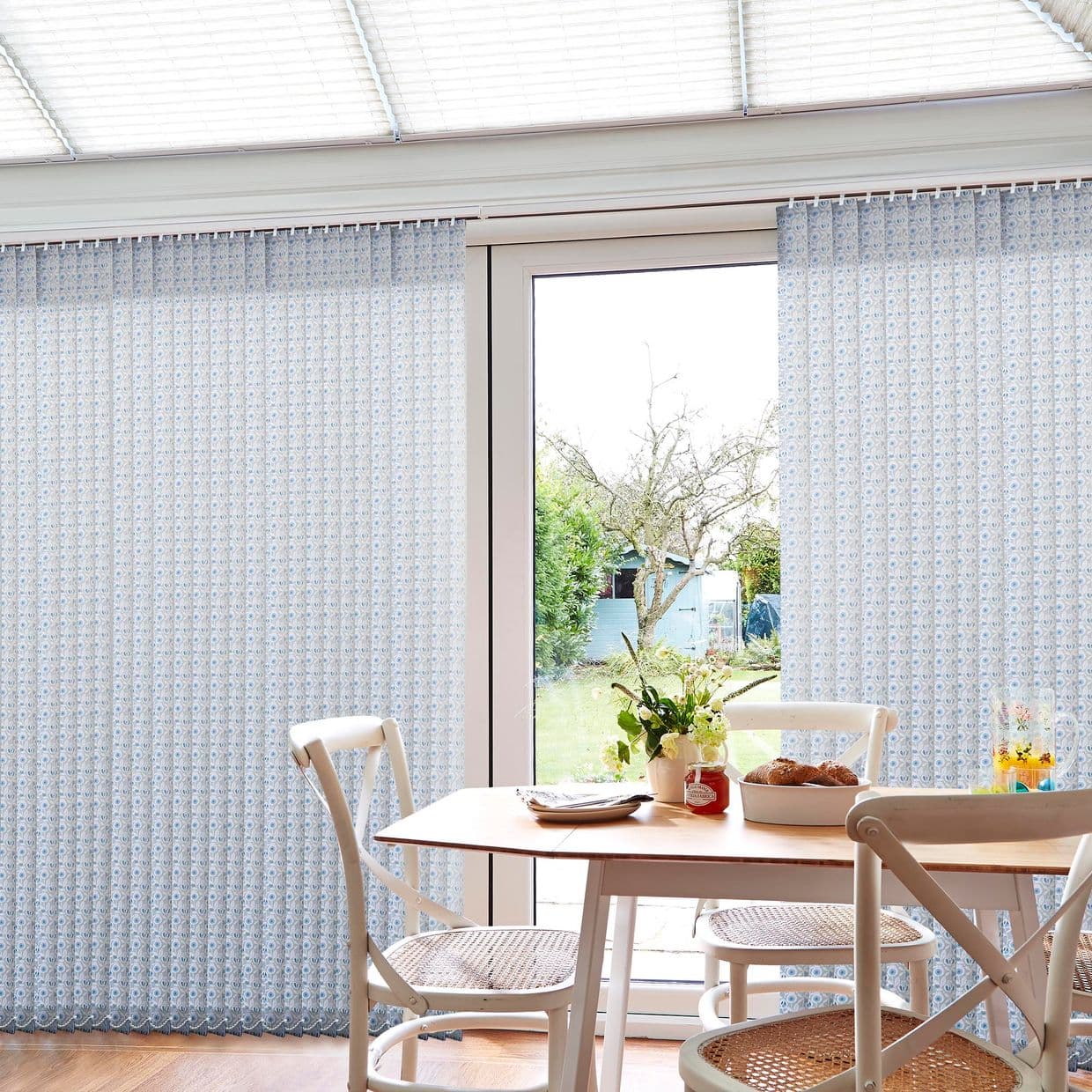 Patterned pale-blue vertical blinds over sliding glass door, partially parted to reveal a sunlit garden; wooden dining table with white chairs, vase of flowers and bread in a bright room.